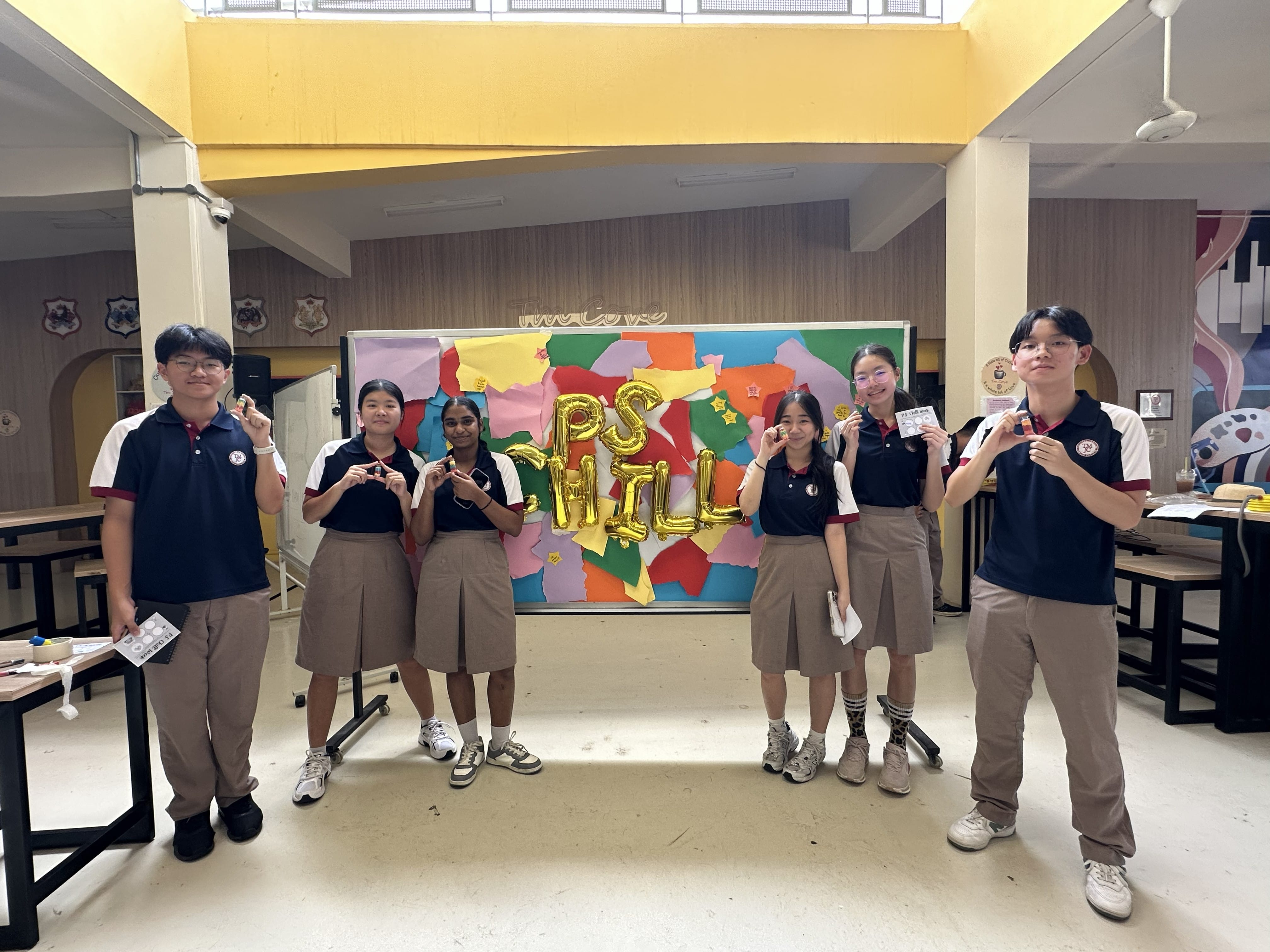 Six students in school uniforms stand in front of the "PS CHILL" balloon backdrop, each proudly holding up their completed sand art bottles.